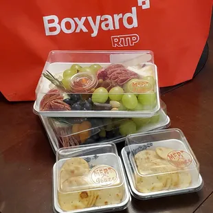 Two of the lunchbox offerings with meat, cheese, fresh and dried fruit, nuts, pickles, chocolate, and honey. With a side of crackers