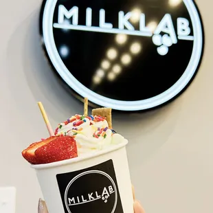 a hand holding a cup of ice cream with strawberries and sprinkles