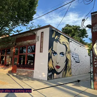 To park, turn into the alley between the woman painting and Riff Raff brick building.