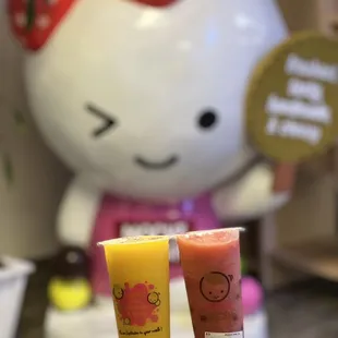 a hand holding two drinks in front of a hello kitty statue