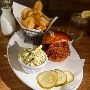 In House Pulled Barbecue Pork Sandwich seated on a Pretzel roll bun with pickles, house-made coleslaw and house-made potatoes chips