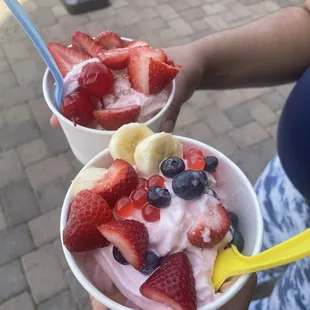 a person holding two bowls of yogurt with strawberries, bananas, and blueberries
