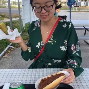 a woman sitting at a table with two hot dogs and a can of soda