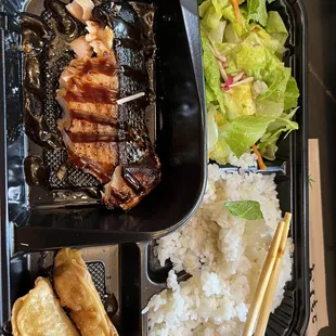 a bento box with rice, meat, salad, and chopsticks