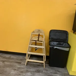 a wooden stool and a black trash can against a yellow wall