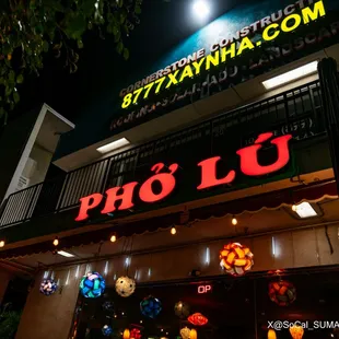 Front of Business: An LED sign that says, &quot;Phở Lú&quot; in glowing red and various decorations on the first floor of a building at night time.