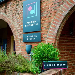 A picture of the Piazza Discepoli sign with logo on a brick wall. The sign is black with white type says Piazza Discepoli Fine Wines &amp; Food.