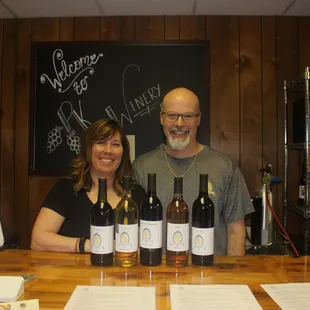 a man and a woman standing behind a counter with bottles of wine