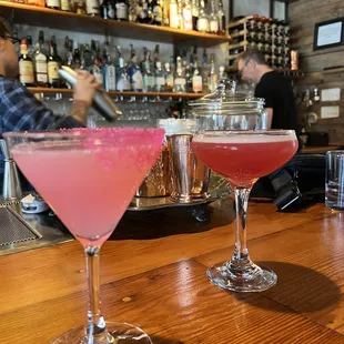 two cocktail glasses on a bar with a bartender in the background