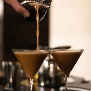 a person pouring a drink into two martini glasses