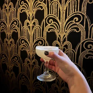 a hand holding a cocktail glass in front of a wallpapered wall