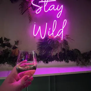 a hand holding a glass of wine in front of a neon sign that says stay wild