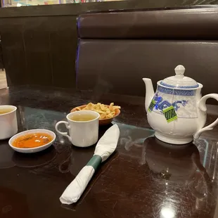 a table with a teapot, cups of soup, and a spoon on it