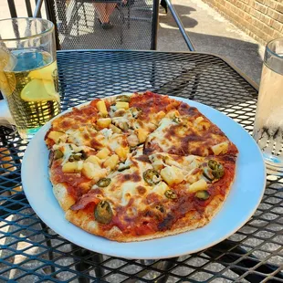 Pineapple cider, Spicy Hawaiian pizza, and a glass of water on an outside table.