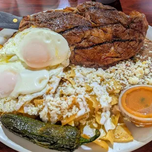 Ribeye Steak Medium Rare, Chilaquiles with Cheese, Eggs Basted, Refried Beans, Home Fried Potatoes, a Roasted Chili and Homemade Salsa.
