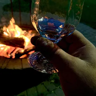 a hand holding a wine glass in front of a fire pit