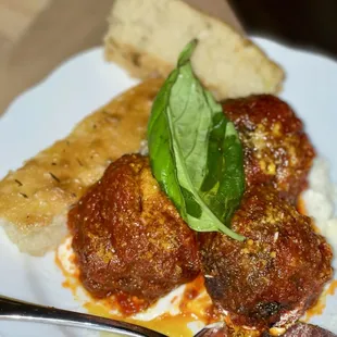 Delicious meatballs (beef/pork) with marinara sauce, melted cheese, a basil garnish, and a slice of herbed bread.
