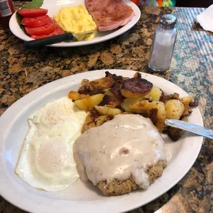 Chicken fried steak hashbrowns and eggs over medium. In the background scrambled eggs ham steak and tomato slices.