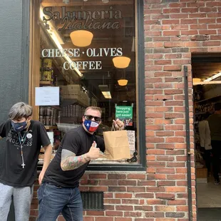 a man in a face mask and a man in a black t - shirt standing in front of a coffee shop