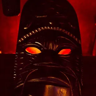 Light flickers behind the eyes of this mask watching over the bar area.