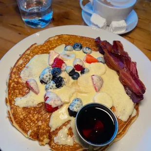 a plate of pancakes with bacon, blueberries, strawberries, and maple syrup