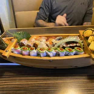 a man sitting at a table in front of a wooden boat filled with sushi