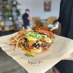 Savage Goods Breakfast Burger: Beef burger, cheddar, bacon, over easy egg, roasted tomato, arugula, and poblano aioli. Served with fries.