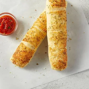 a pair of breadsticks on a piece of paper with a small bowl of ketchup