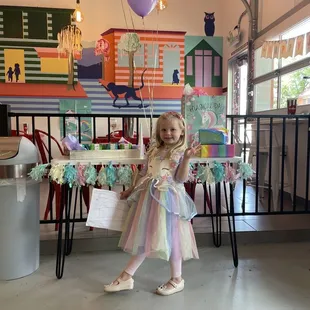 a little girl in a dress standing in front of a table with balloons