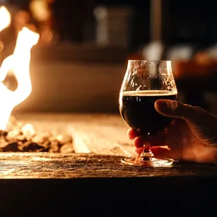a person holding a glass of beer in front of a fire