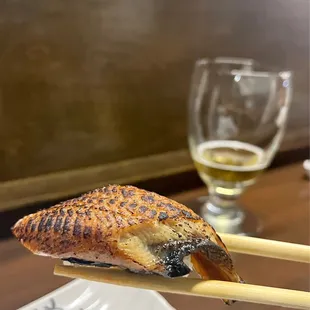 a piece of fish on chopsticks with a glass of beer in the background