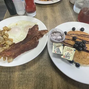 two plates of pancakes, bacon, blueberries, and eggs