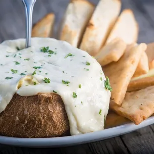 This creamy dip blends roasted garlic &amp; artichoke hearts with melted cheese, served in a sourdough bread bowl w/ crispy pita &amp; veggies!