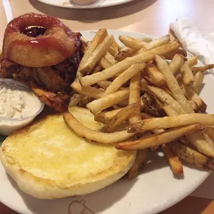Pulled pork sandwich with french fries and coleslaw. Fried onion ring is on top of the sandwich.