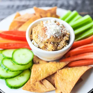 a white plate with a bowl of hummusle, carrots, cucumbers, and crackers