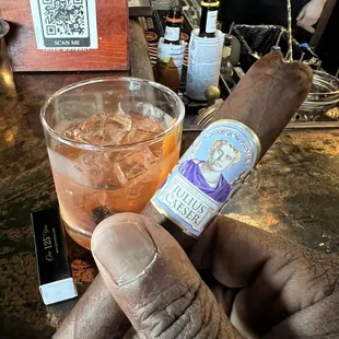 a hand holding a cigar and a glass of whiskey