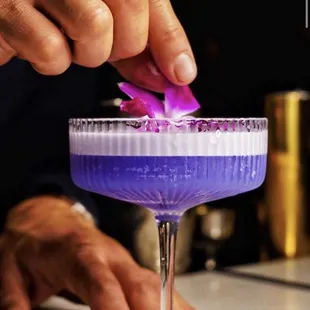 a close up of a person&apos;s hand placing a purple flower into a cocktail glass