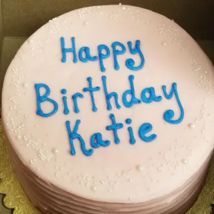 A round, gluten-free birthday cake with pale pink icing &amp; white edible &quot;pearls.&quot; The words &quot;Happy Birthday Katie&quot; are in the middle in blue.