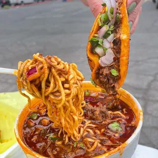 a hand holding a taco over a bowl of noodles