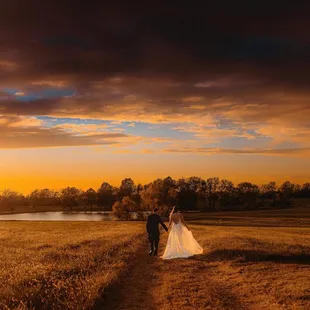 a bride and groom walking in a field at sunset