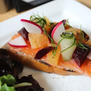 Gravlax (cured salmon) on top of a grilled baguette with dill labneh, beets, pickles, and a petite salad on the side.
