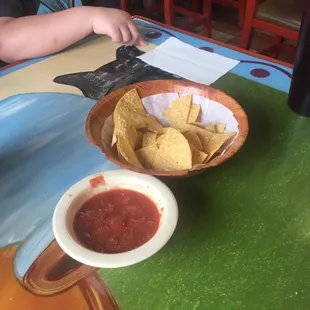 a little boy sitting at a table with a bowl of chips and a bowl of salsa