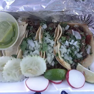 Tacos asadas in corn tortillas with cilantro, onion, lime, radishes, cucumber, and salsa verde.