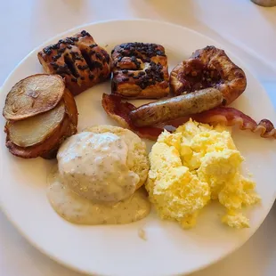 Breakfast buffet plate: Biscuit and gravy, scrambled eggs, potatoes, sausage, bacon, pastries.