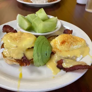 a plate of eggs benedict with bacon and a bowl of fruit