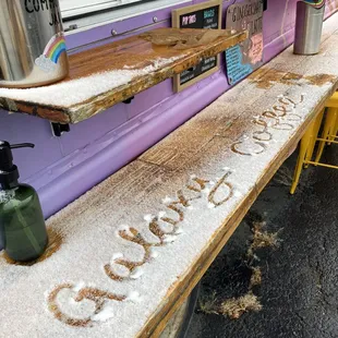 The words "Galaxy Coffee" written in the snow that is lightly dusted on the wooden counter attached to The Galaxy Coffee truck