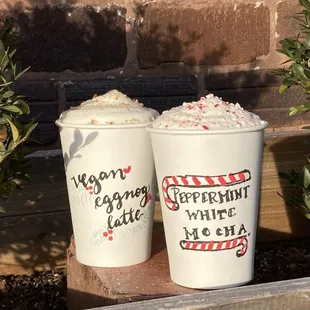 Two lattes set in front of a brick wall, both with cloud foam piled on top and candy crunch on one, and cookie crunch on the other.