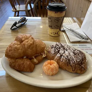 The English breakfast Sandwich on a fresh croissant (left). Chocolate-Almond croissant (right), a little tangerine, and latte.