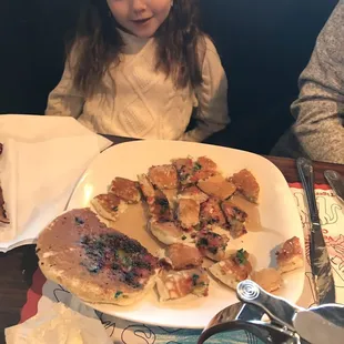 a little girl sitting at a table with a plate of pancakes