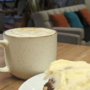 Latte in a mug with iced cinnamon roll on a plate, partial windmill and couch in the background
